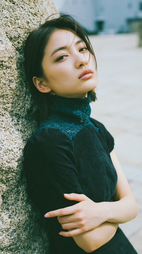 A young Japanese woman with short black hair leans against a rock, looking towards the camera with a serene expression.