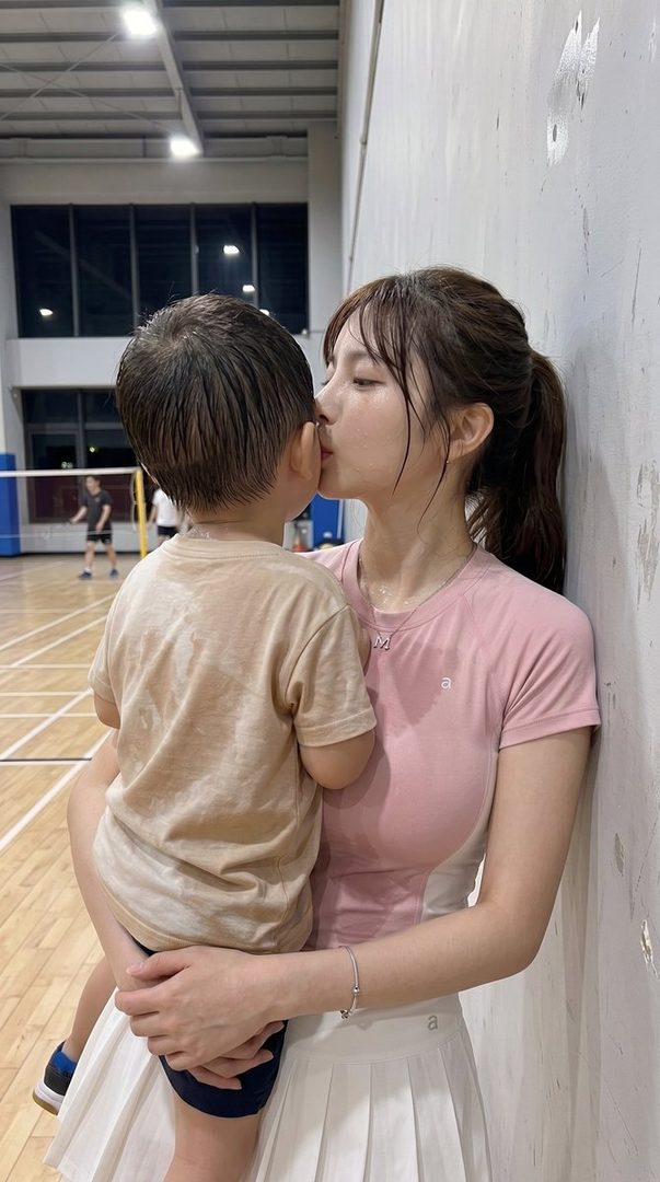 A young woman in a pink t-shirt and white skirt holds a young child, leaning in to kiss him, in an indoor sports facility.