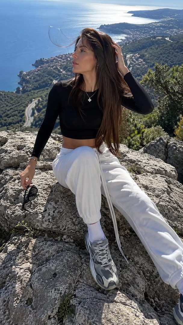 A woman with long brown hair sits on a rock overlooking a coastal landscape, wearing a black crop top and white joggers.
