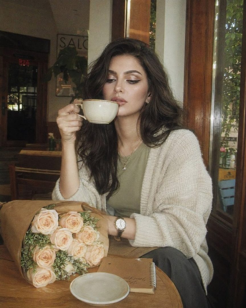 A woman with long dark hair sits at a cafe table, holding a coffee cup and looking thoughtfully downwards, with a bouquet of roses beside her.