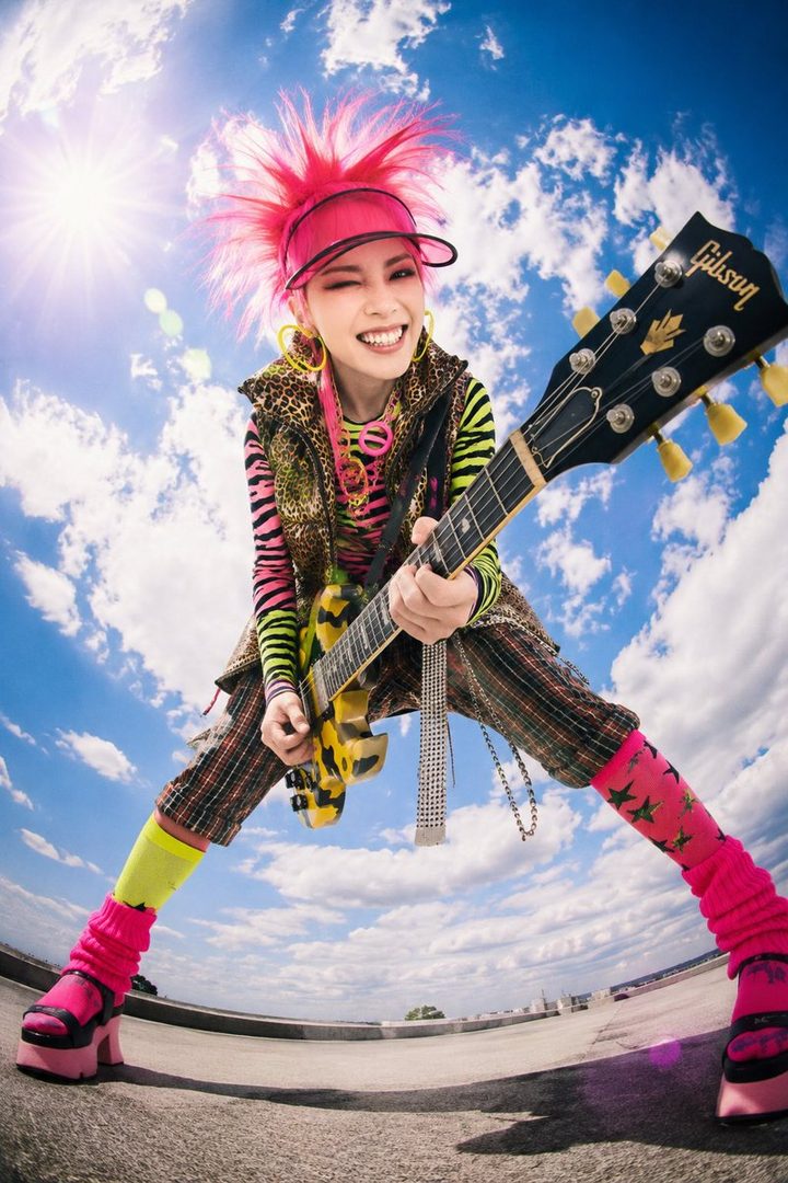 A young woman with bright pink spiky hair and a vibrant, punk-inspired outfit, holding a guitar and smiling playfully against a bright blue sky.