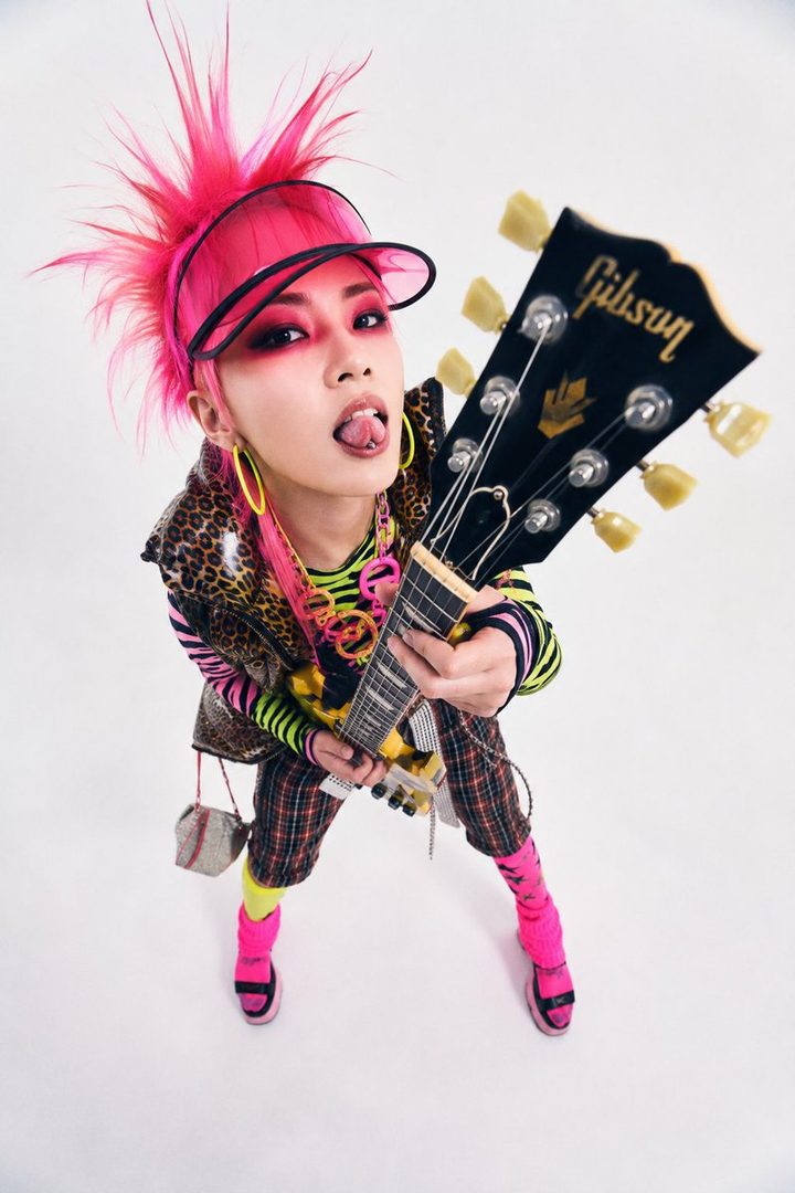 A young woman with bright pink spiky hair and punk-inspired makeup, wearing a leopard print vest over a striped shirt and plaid pants, plays a guitar.