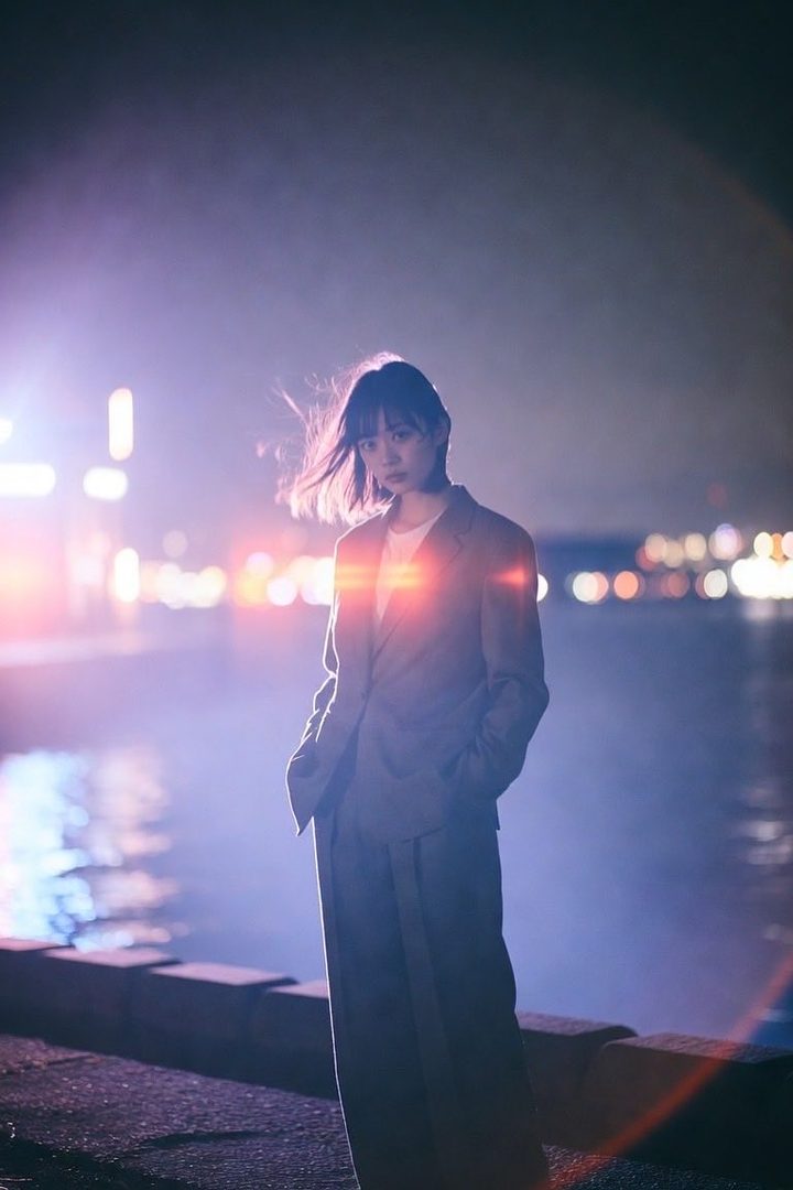 A young woman in a suit stands by the water at night, with city lights blurred in the background.