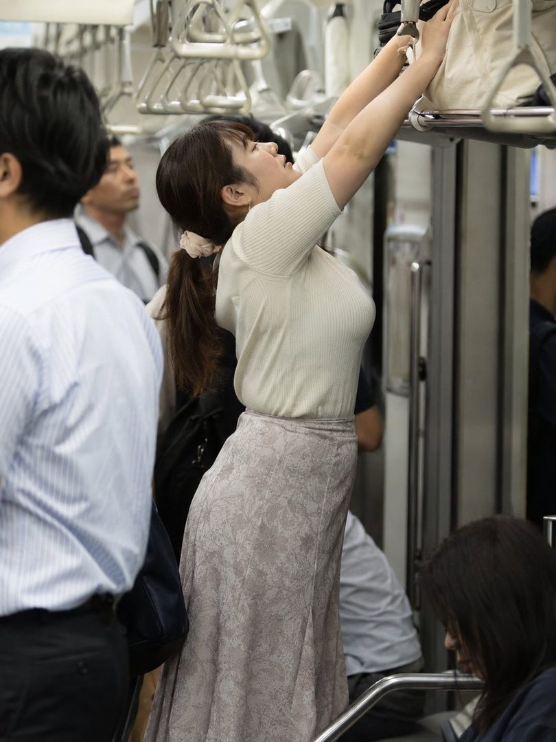 A young woman with brown hair in a ponytail, wearing a white top and a patterned skirt, stands on a crowded train, reaching for a handle.