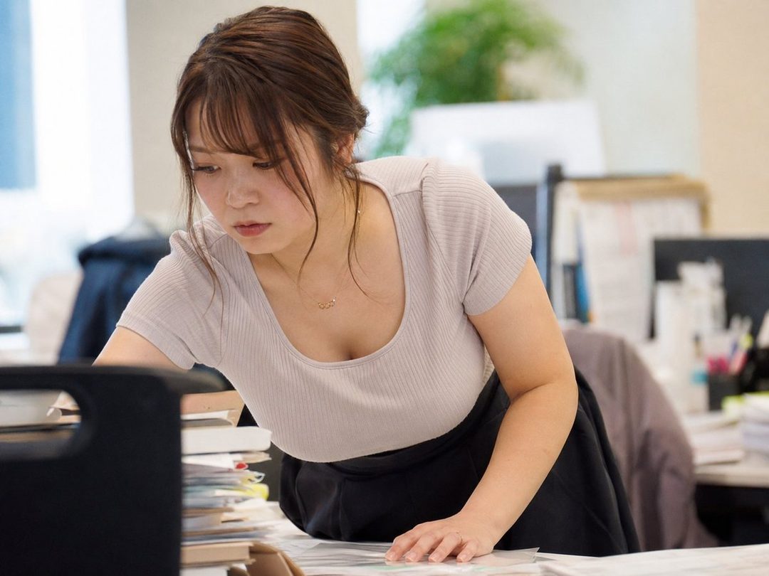 A young East Asian woman with brown hair and bangs, wearing a light-colored ribbed top and a black skirt, leans over a desk in an office setting, looking down at papers.