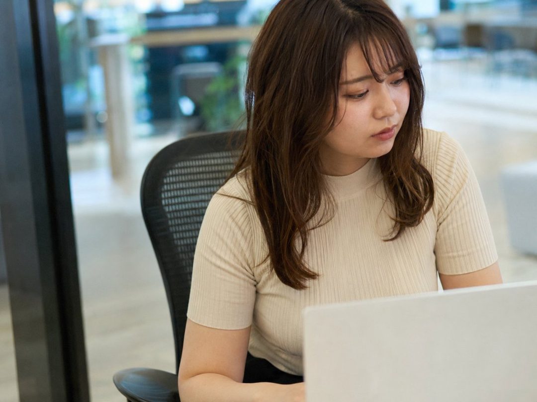 A young East Asian woman with long brown hair sits at a desk in a modern office, looking down at her laptop.