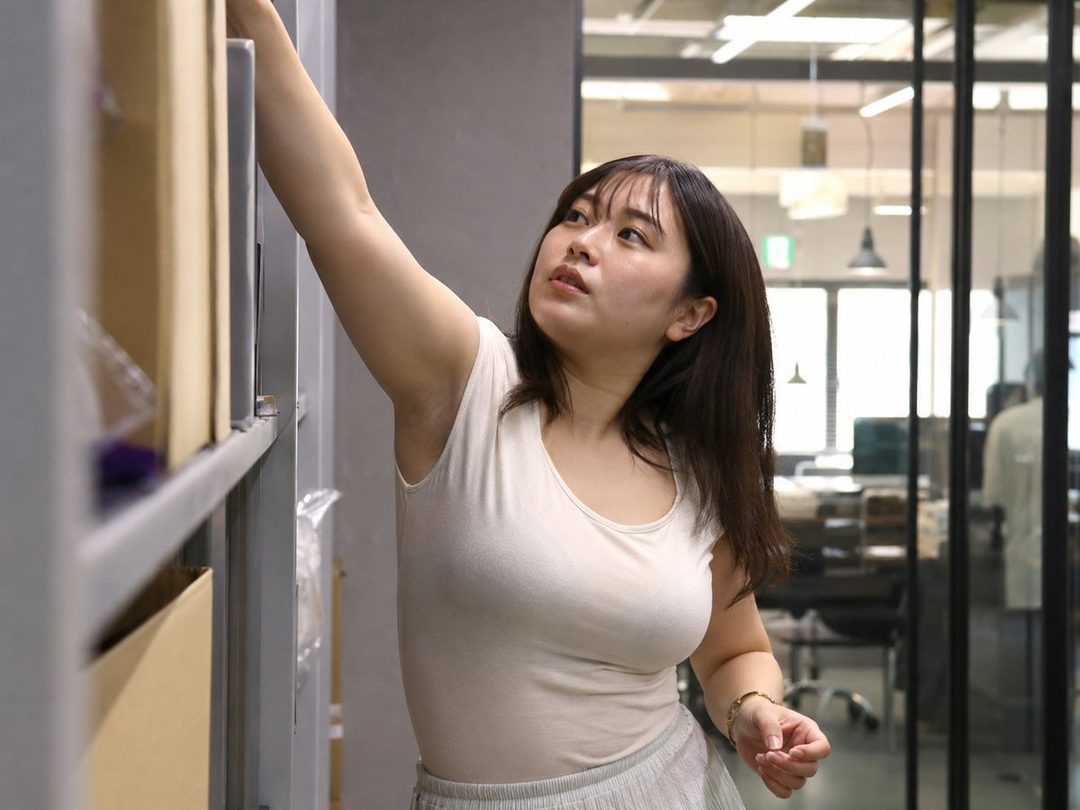 A young East Asian woman with long dark hair reaches for a box on a shelf in an office setting.