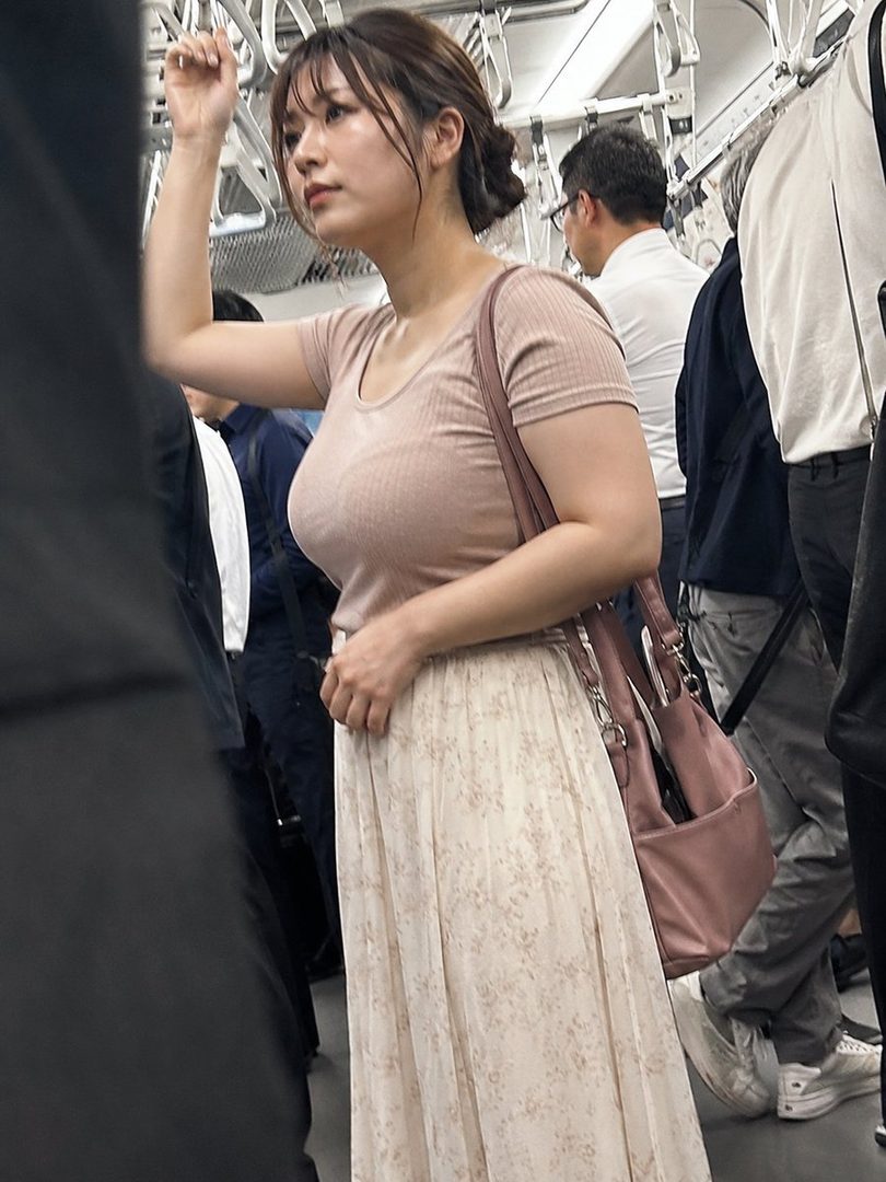 A young woman with her hair tied up in a bun, wearing a light pink t-shirt and a floral print skirt, stands on a crowded train, holding onto the overhead handrail.