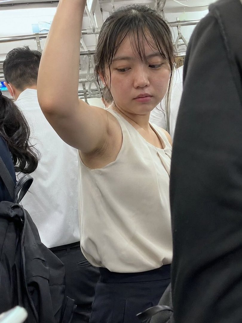 A young East Asian woman with bangs and dark hair, wearing a light-colored sleeveless top and dark pants, is holding onto a pole on a crowded train during the daytime.