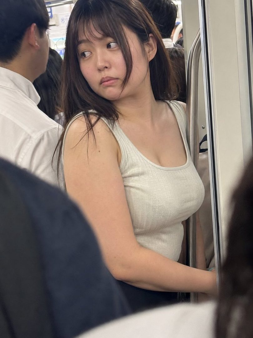 A young Japanese woman with long brown hair and bangs, wearing a light-colored tank top, looks away while riding a crowded train during the summer.
