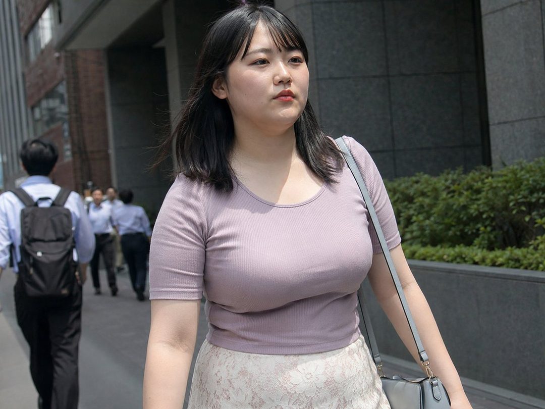 A young East Asian woman with black hair, wearing a light purple ribbed t-shirt and a white lace skirt, walks down a city street on a sunny day.