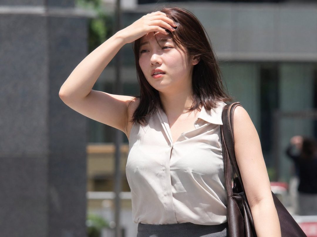 A young East Asian woman with brown hair, wearing a light-colored sleeveless blouse and a grey skirt, shields her eyes from the sun with her hand while walking outdoors.