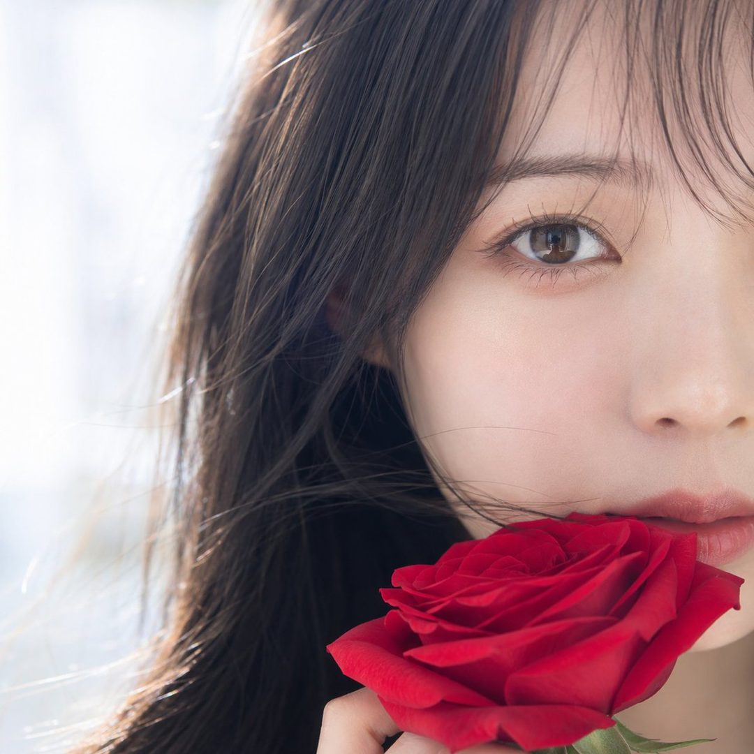 Close-up portrait of a young East Asian woman with long dark hair holding a red rose to her lips, soft lighting.