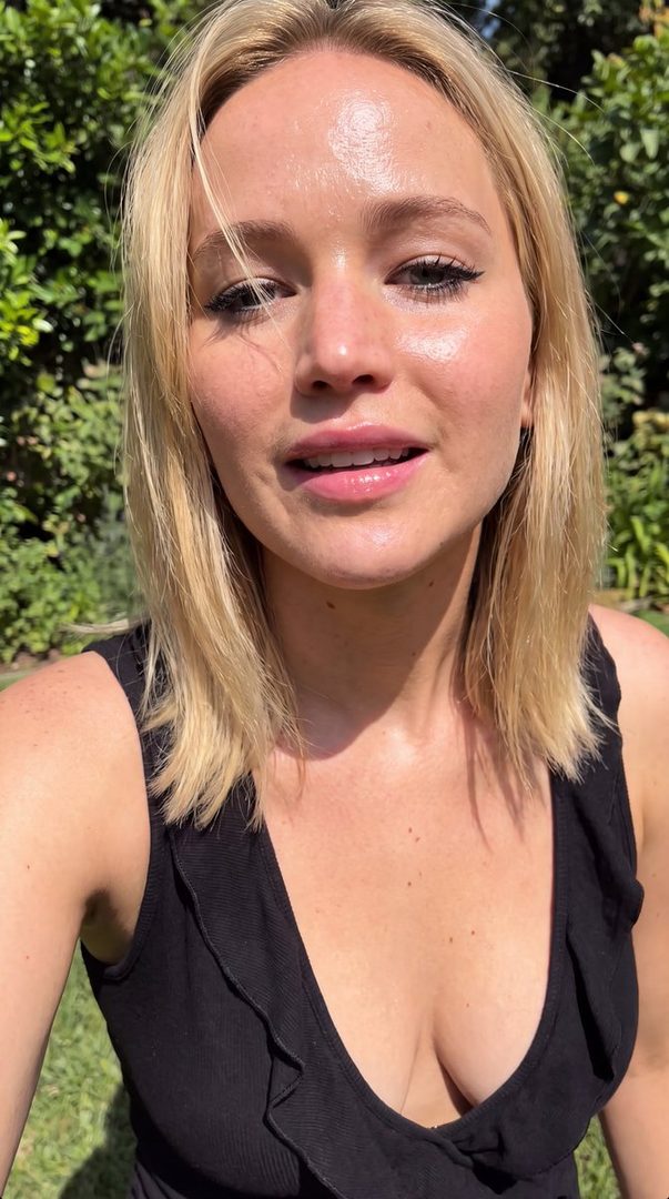 A blonde woman with fair skin and blue eyes, wearing a black tank top, smiles at the camera in an outdoor setting with green foliage in the background.