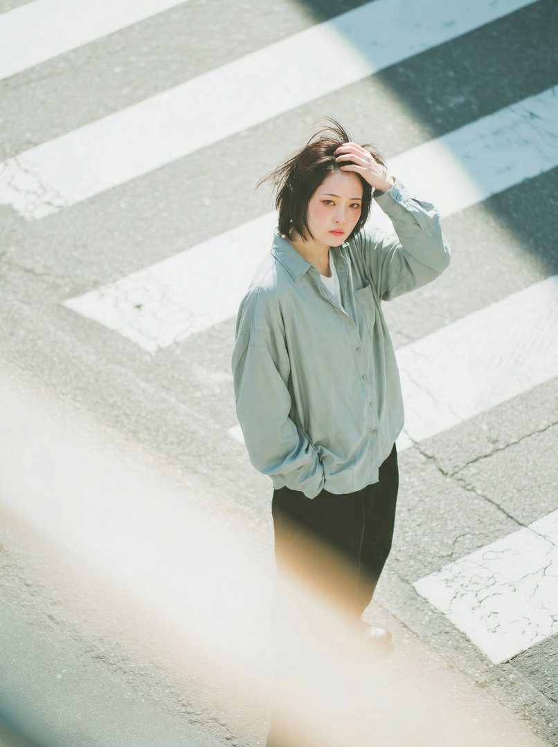 A young woman with short black hair and light makeup stands on a crosswalk, touching her hair.
