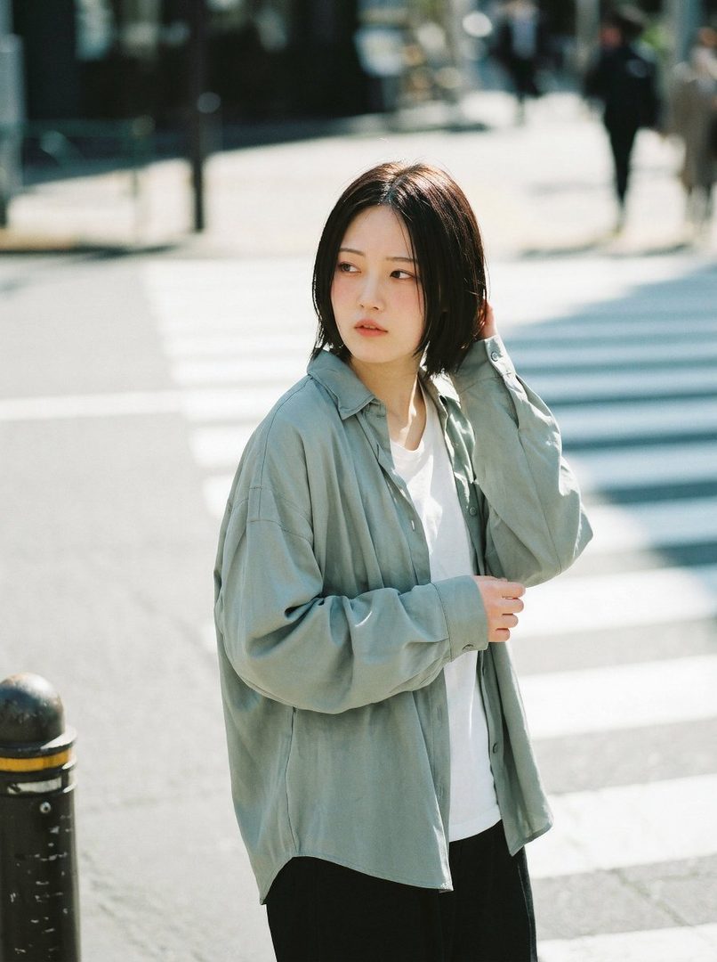 A young East Asian woman with short black hair stands on a city street, looking to the side.