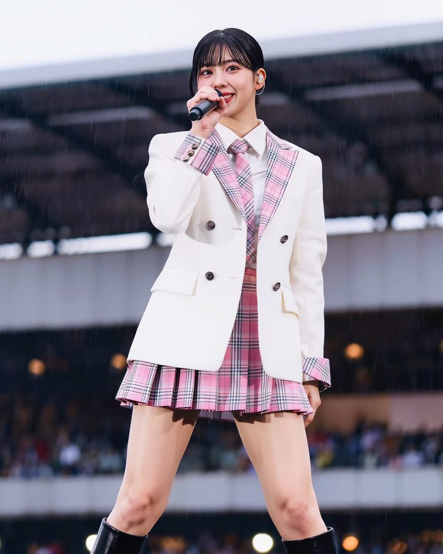 A young East Asian woman in a white blazer, pink plaid tie, and matching pleated skirt, holding a microphone and singing on stage.
