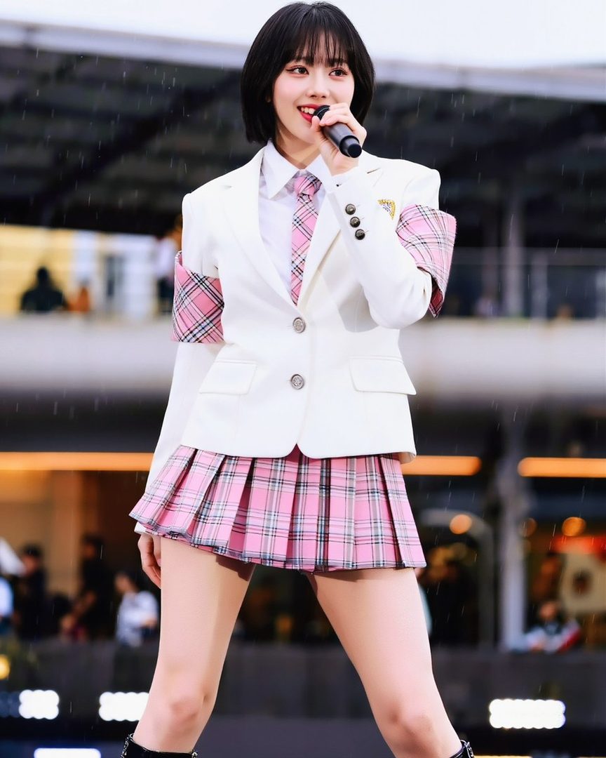 A young East Asian woman with short black hair, wearing a white blazer, pink plaid tie, and pink plaid pleated skirt, holding a microphone and singing on stage.