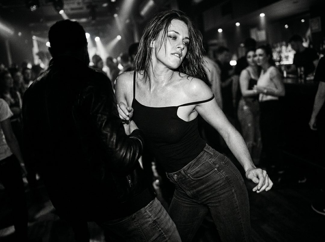 A woman dancing intensely in a crowded nightclub, sweat on her skin, wearing a black tank top and jeans.