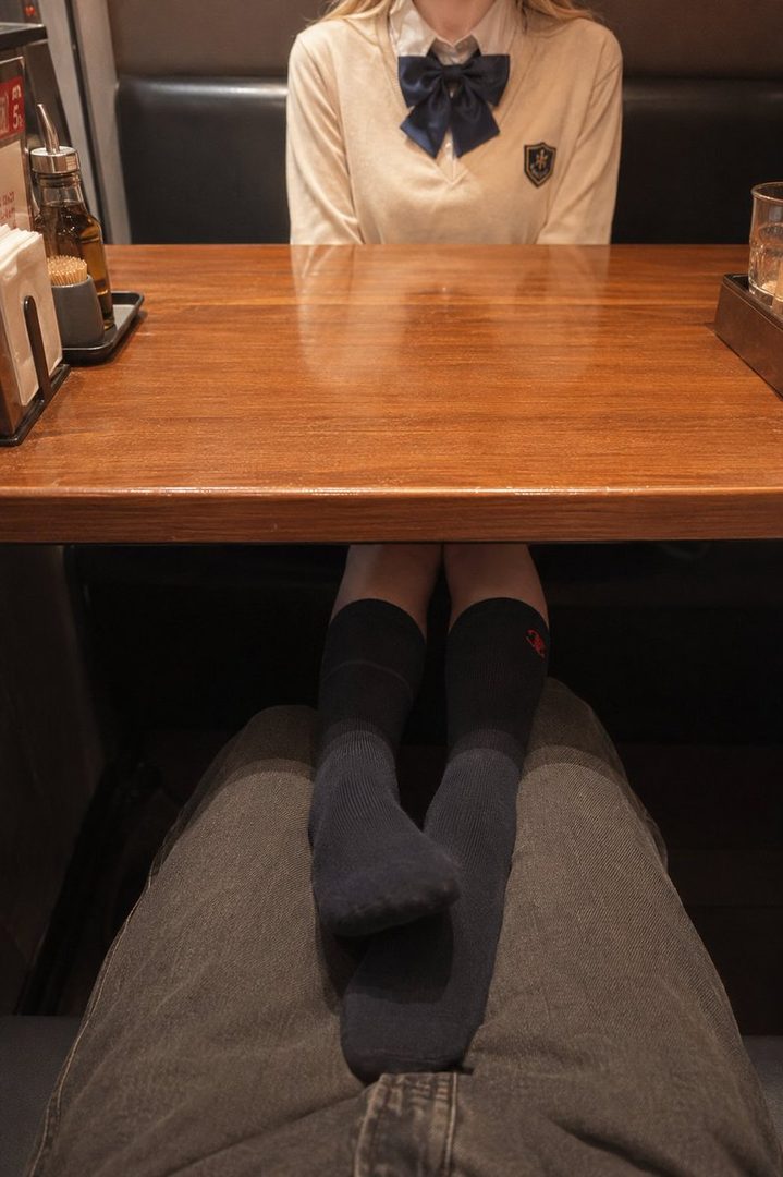 A young woman wearing a school uniform with a blue bow tie sits at a wooden table, her legs crossed and wearing knee-high socks.