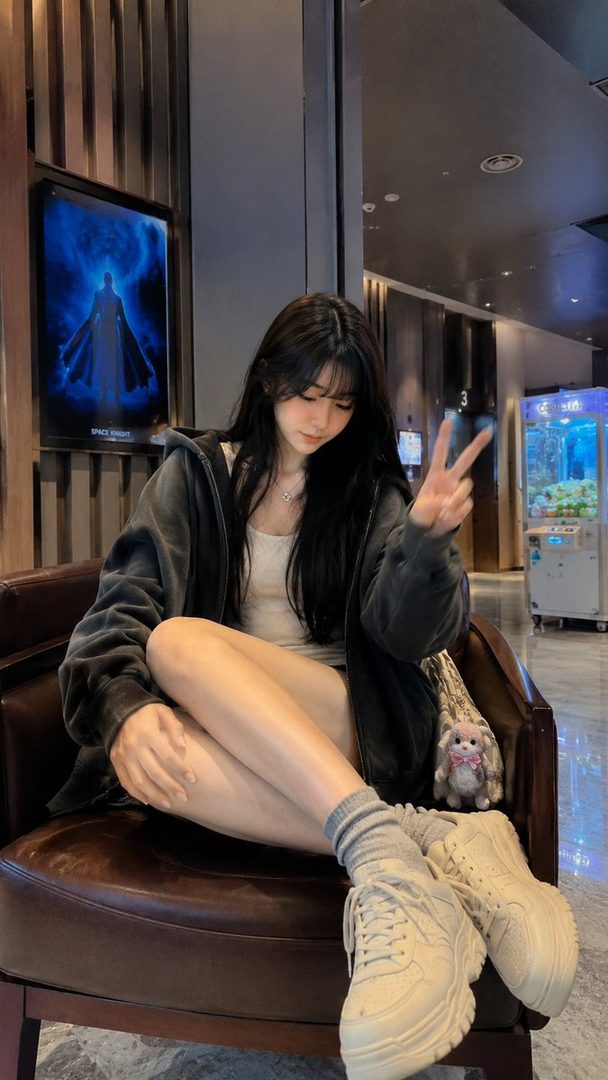 A young woman with long black hair sits in a chair, wearing a grey zip-up hoodie over a white tank top and grey socks with chunky sneakers. She is making a peace sign with her fingers and looking down.