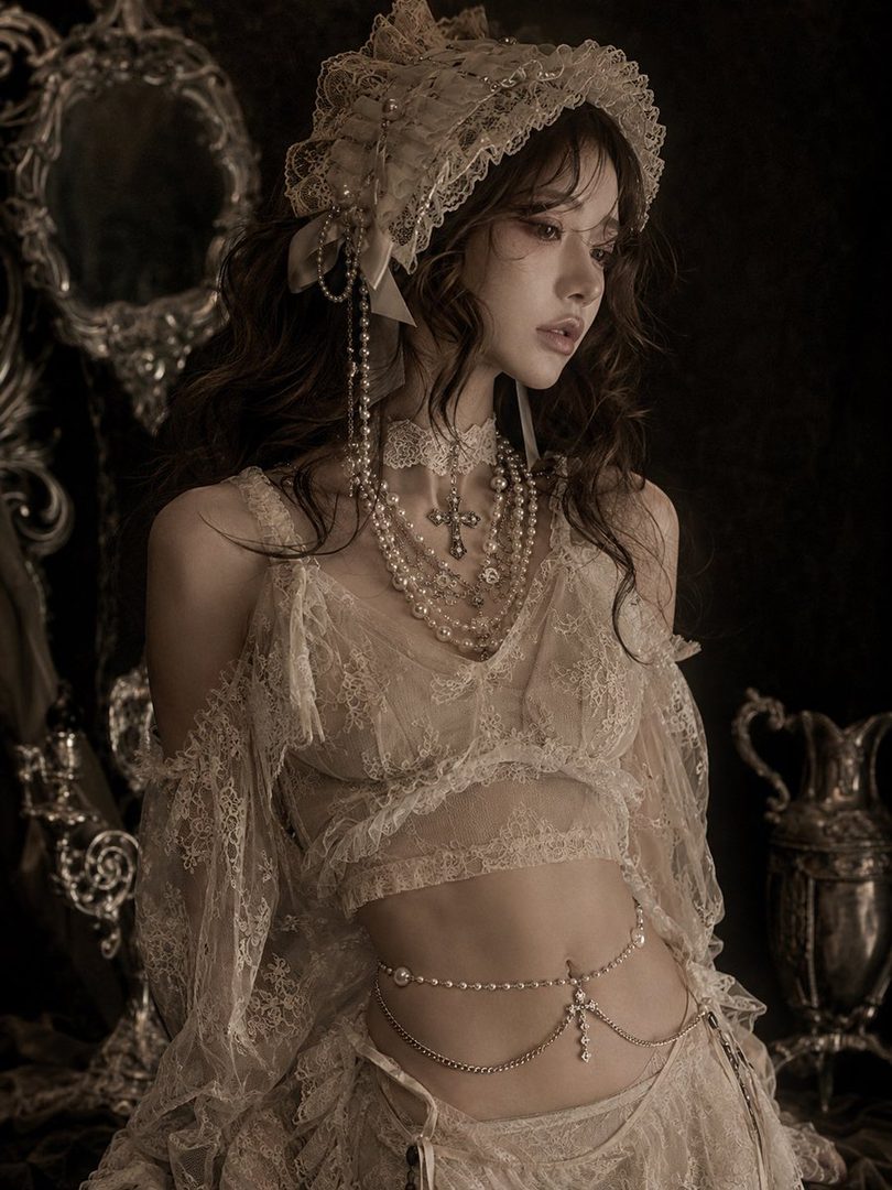 A young woman with long brown hair, wearing a vintage-inspired lace outfit and elaborate jewelry, poses elegantly indoors.