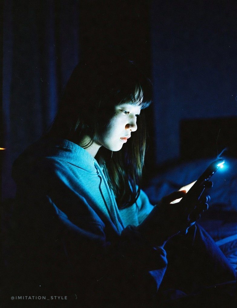 A young East Asian woman with long black hair, illuminated by the blue light of her smartphone, looking down at the screen in a dimly lit room.