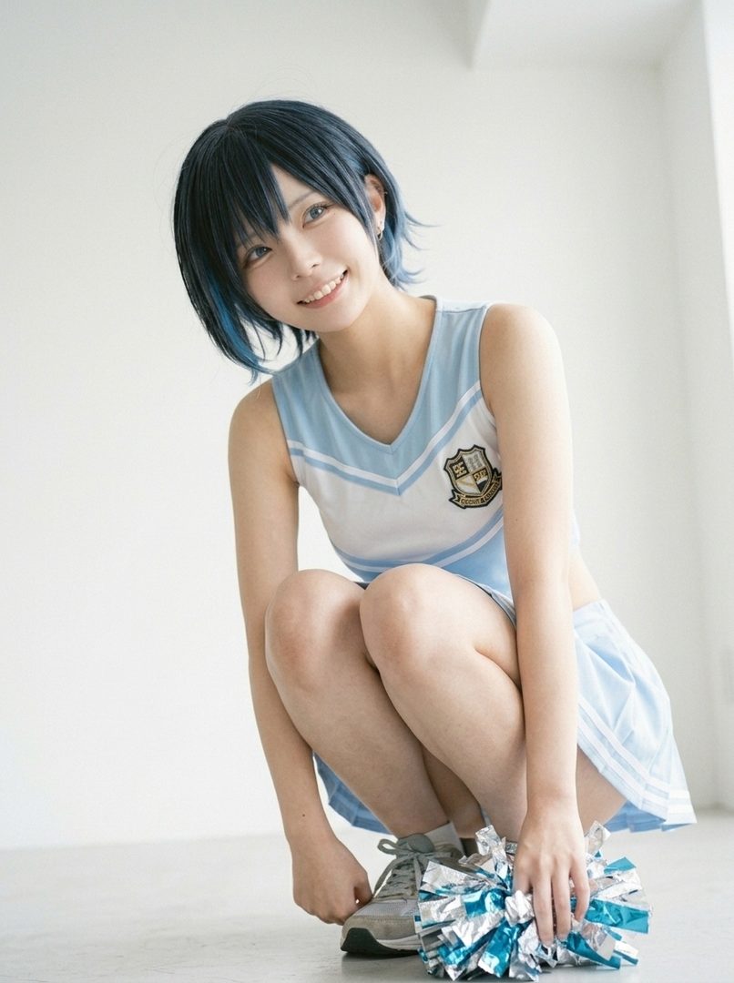A young woman with short blue-black hair, wearing a light blue and white cheerleader outfit, squats down holding a pom-pom.