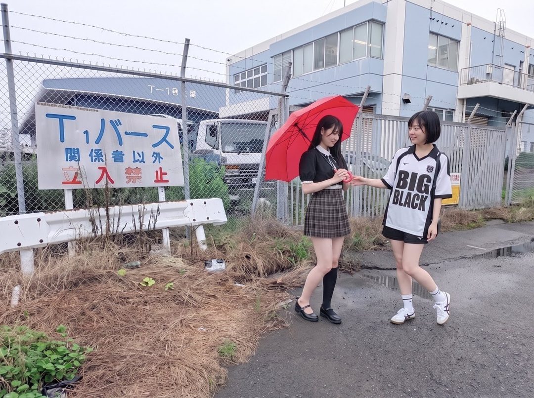 Two young women, one holding a red umbrella, stand outside a building with a fence and a 'No Entry' sign. The woman on the left wears a black top and plaid skirt, while the woman on the right wears a white and black jersey.