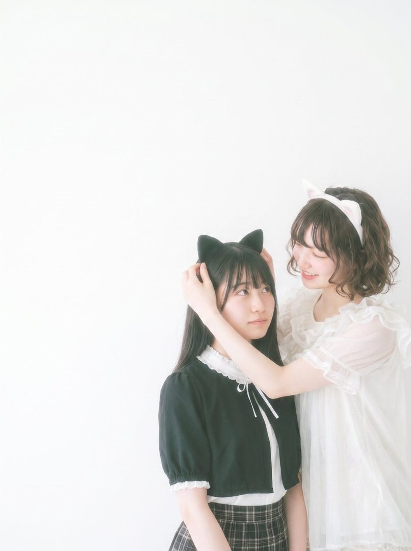 Two young East Asian women wearing cat ear headbands, one adjusting the other's headband in a studio setting.