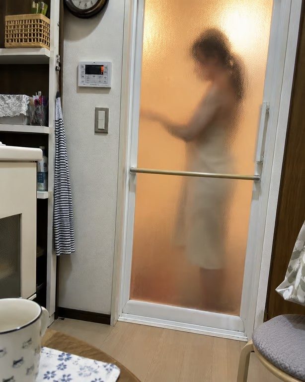 A woman in a towel is seen through a frosted glass shower door, with warm light illuminating the scene.