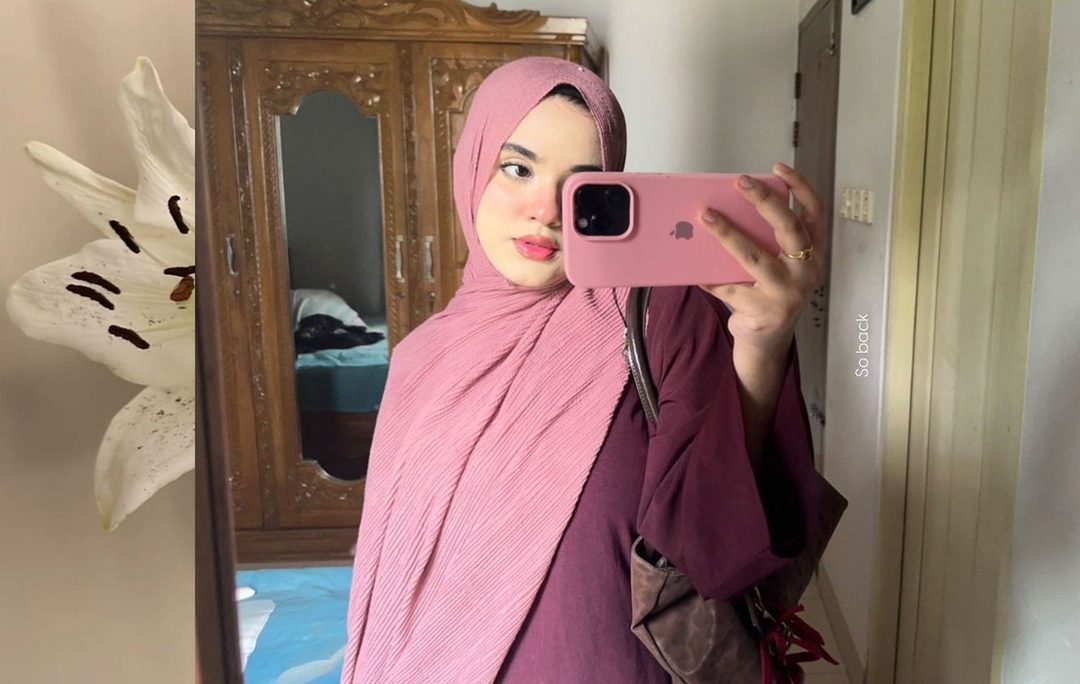 A young woman wearing a pink hijab and a maroon top takes a selfie in a mirror, holding a pink phone.