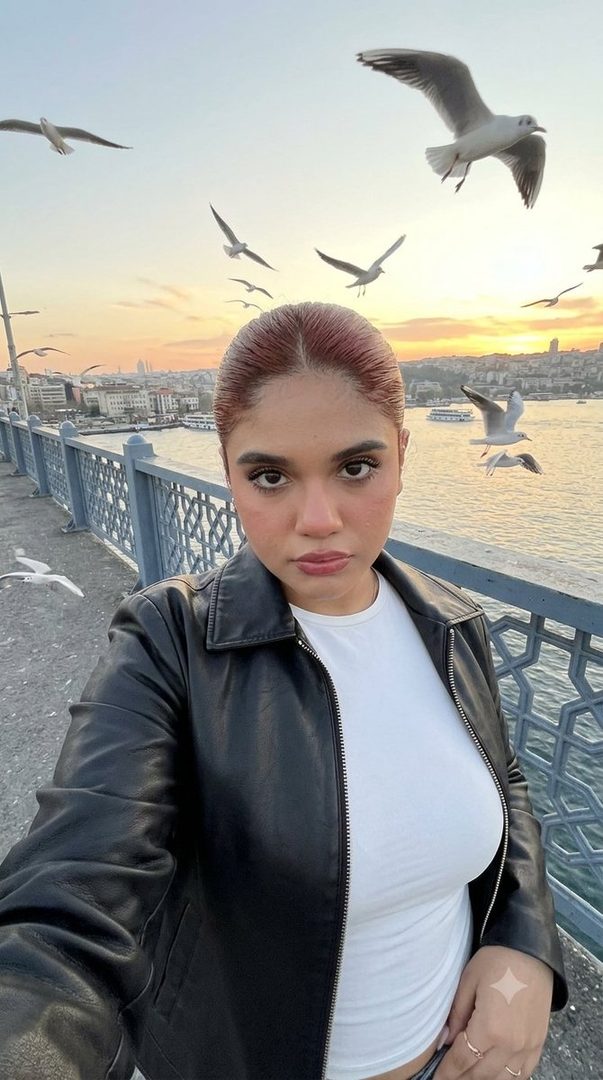 A young woman with red hair pulled back takes a selfie on a bridge with seagulls flying around her at sunset.
