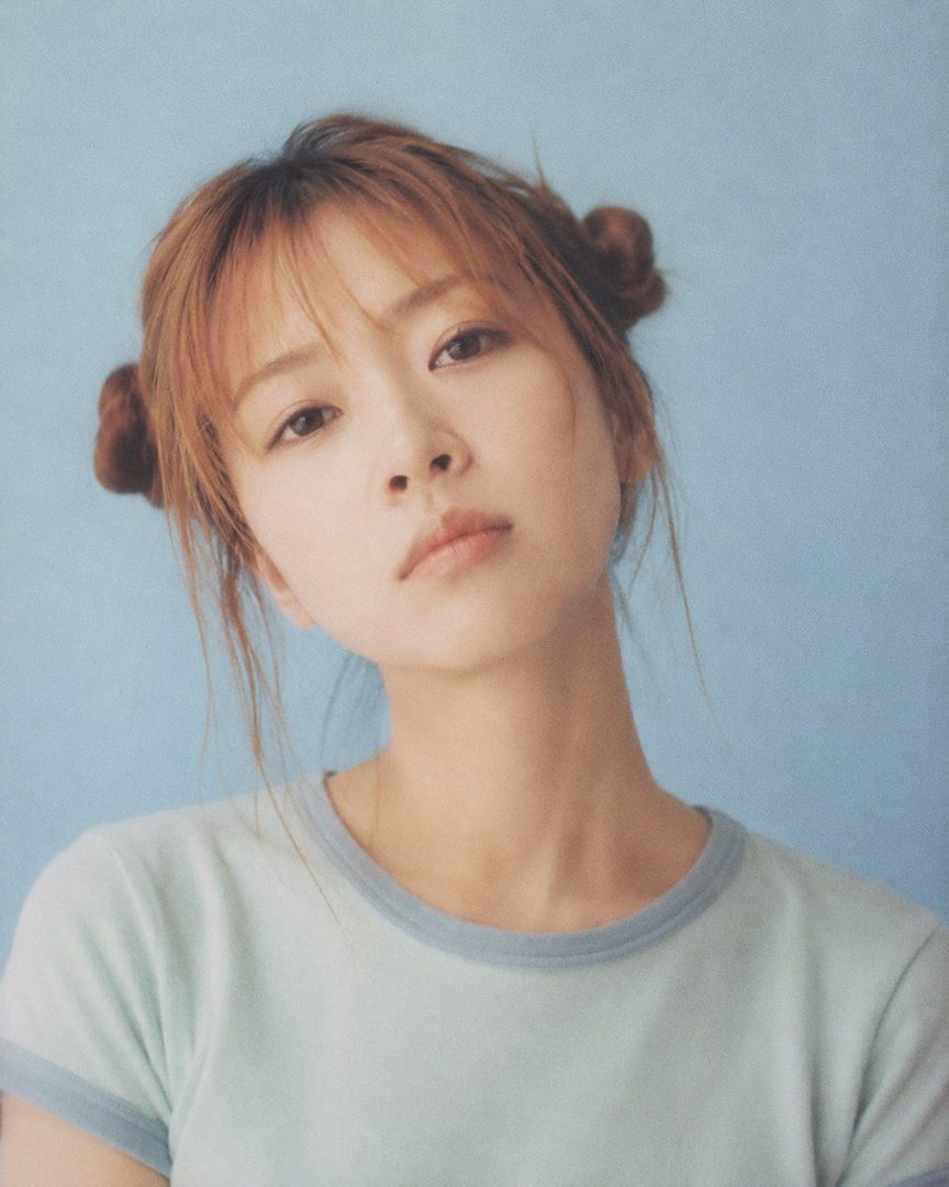 A young woman with brown hair styled in twin buns and bangs, wearing a light blue t-shirt, poses against a light blue background.