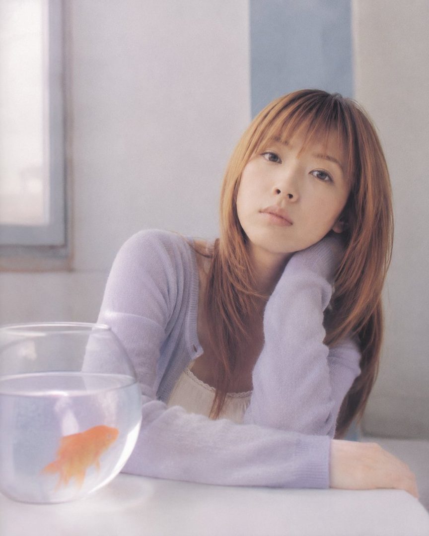 A young Japanese woman with long brown hair and bangs, wearing a light purple cardigan over a white camisole, rests her head on her hand next to a fishbowl containing a goldfish. The soft, natural light creates a dreamy and serene atmosphere.