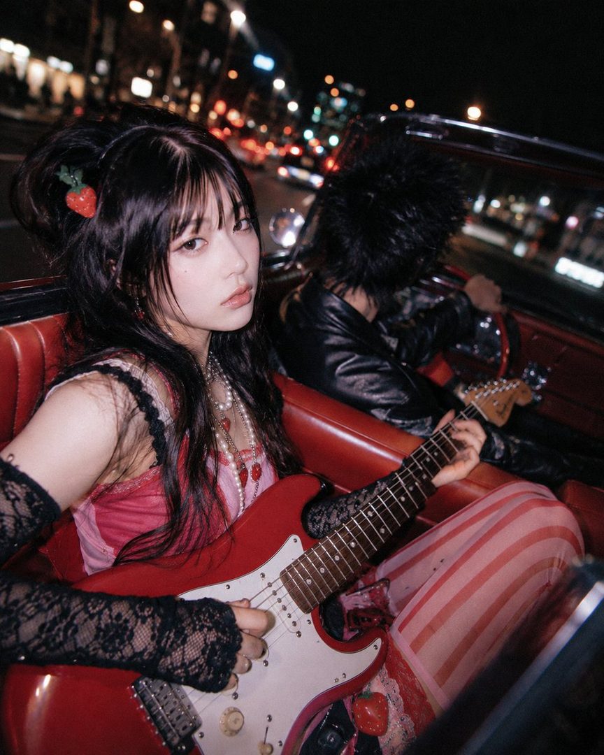 A young woman with black hair and bangs, wearing a pink dress and lace gloves, plays a red electric guitar in the back of a car at night, with city lights in the background.
