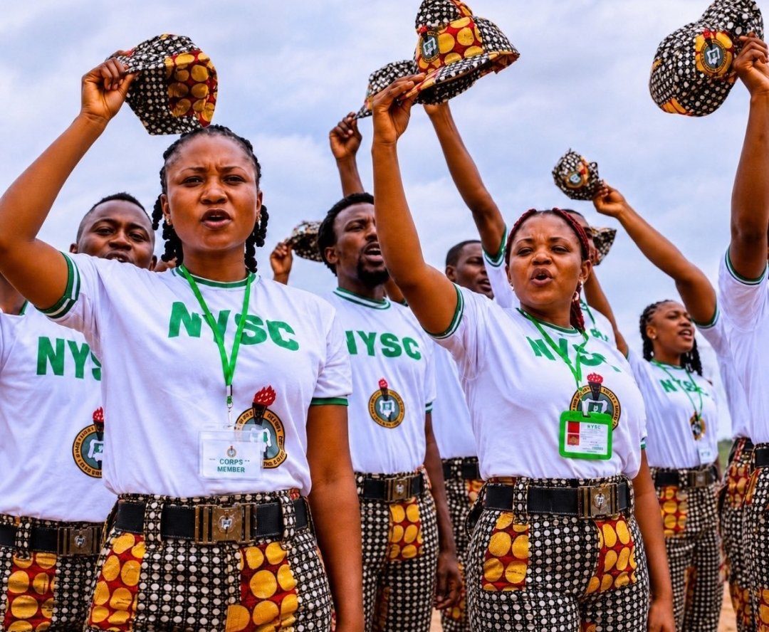 A group of young adults in white t-shirts and patterned trousers are raising their hats in an outdoor setting.