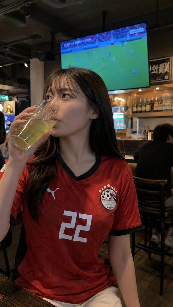 A young woman wearing an Egyptian national football team jersey drinks from a glass while watching a soccer match on TV.