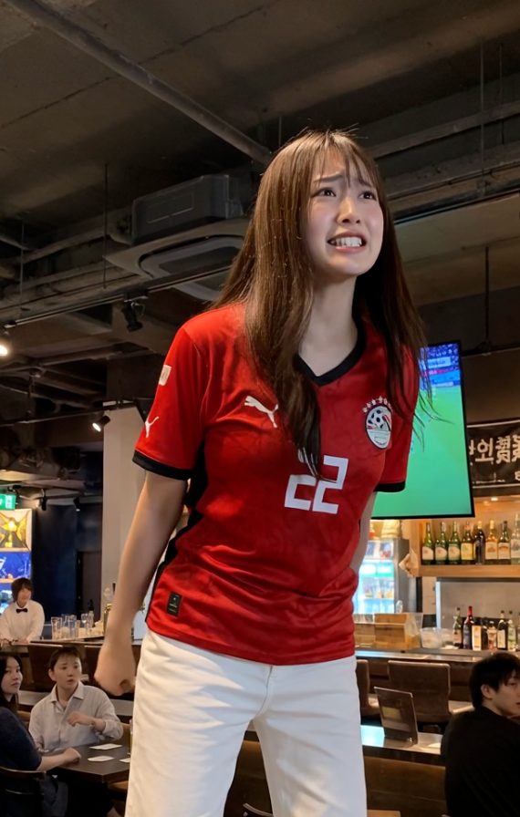 A young woman wearing an Egyptian football jersey and white pants, with a distressed expression, indoors.