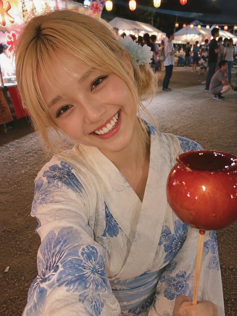 A young woman with blonde hair tied up, wearing a blue and white floral yukata, smiles brightly while holding a candied apple at a summer festival.