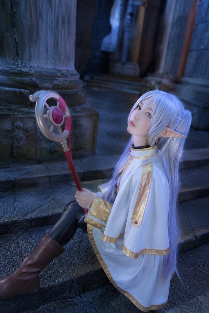 A young woman with long, light purple hair and elf ears, dressed in a white and gold fantasy-style outfit, holding a staff, sits on stone steps outdoors.