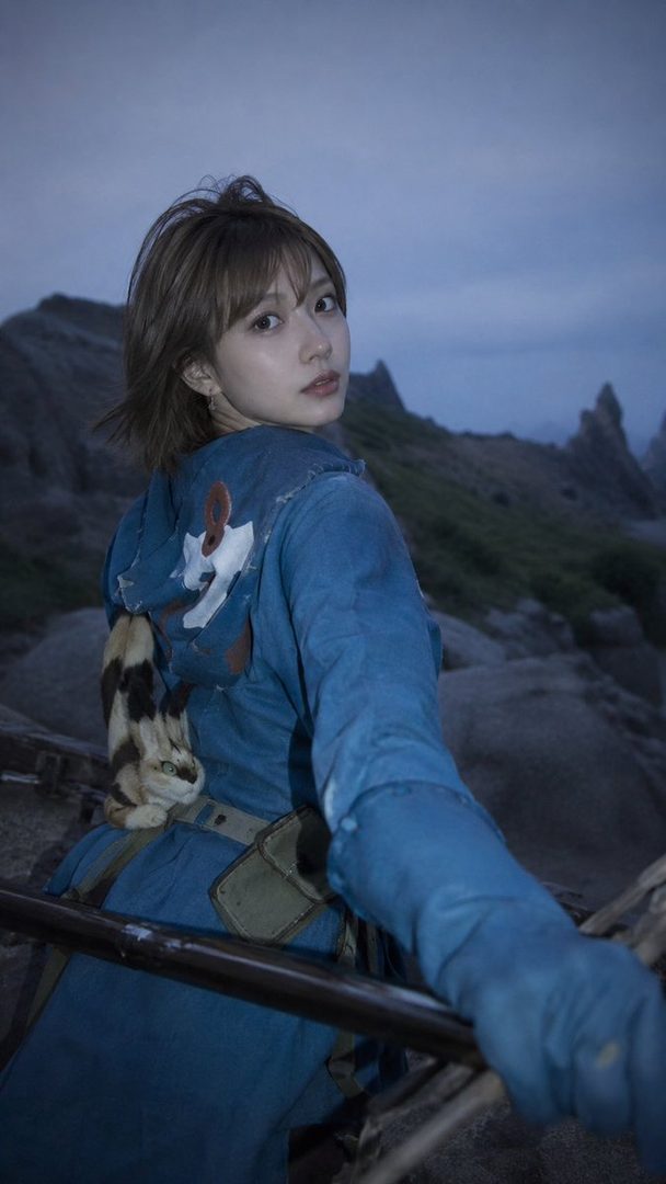 A young woman with short brown hair looks over her shoulder, wearing a blue hooded jacket with a cat accessory on her back, standing outdoors in a rocky, mountainous landscape.