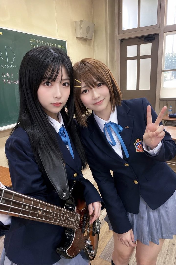 Two young women in school uniforms, one holding a bass guitar, posing for a photo in a classroom setting.