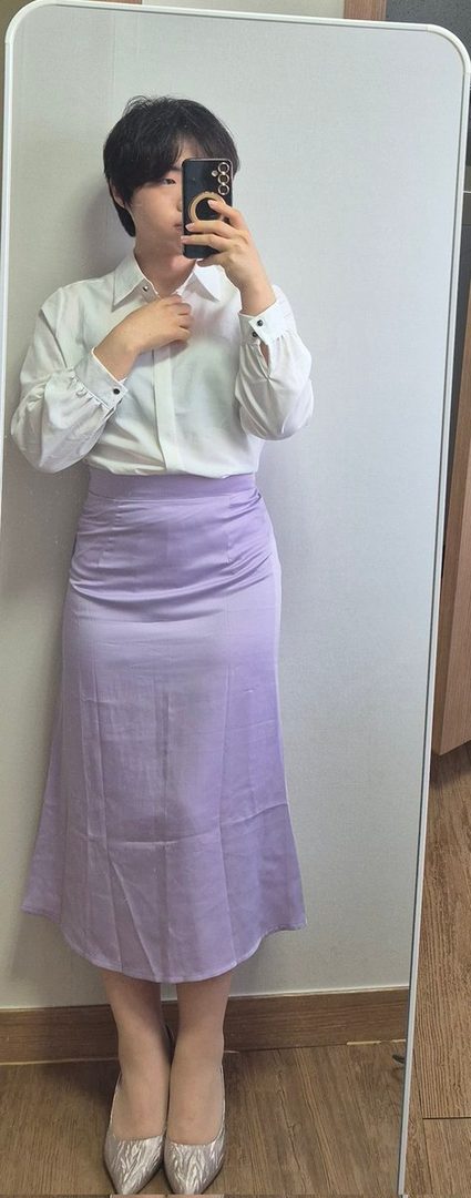 A person with short dark hair takes a mirror selfie, wearing a white blouse and a long lavender skirt, with their face partially obscured by a phone.