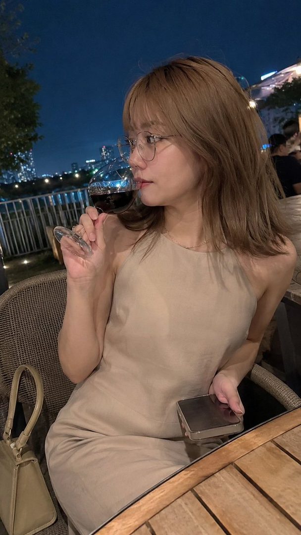 A young woman with brown hair and glasses drinks red wine on a terrace at night, with city lights in the background.