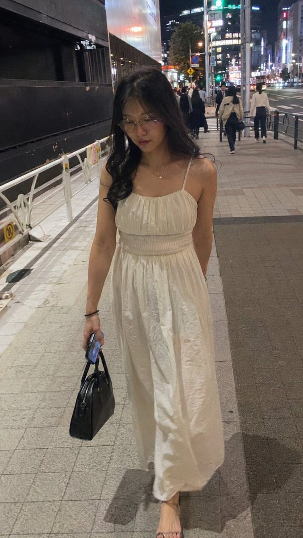 A young woman with long dark hair and glasses walks down a city street at night, wearing a cream-colored dress and carrying a black handbag.