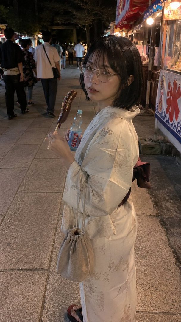 A young woman in a yukata and glasses holds a chocolate-covered banana and a bottle of Ramune soda at a night festival.