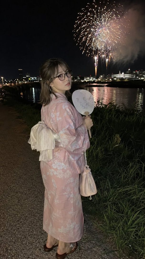 A young woman in a pink floral yukata stands outdoors at night, holding a fan, with fireworks exploding in the sky behind her.