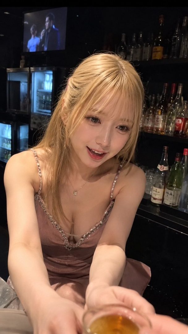 A young woman with blonde hair smiles at the camera while holding a drink in a bar setting.