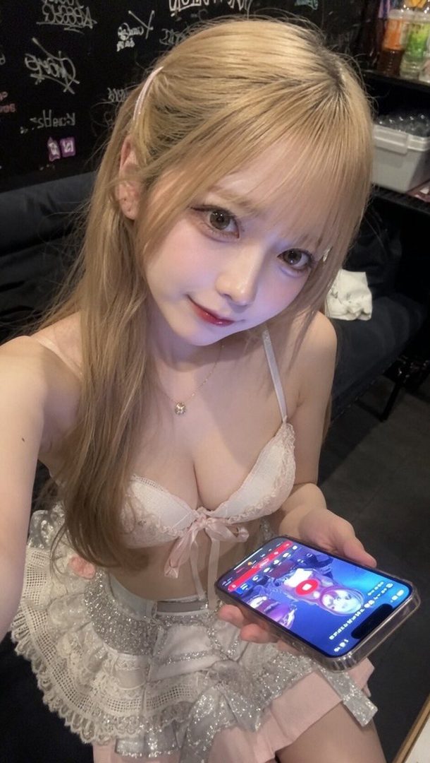 A young Japanese woman with long blonde hair and bangs, wearing a white lace bra and a sparkly skirt, holds up a smartphone showing a game. She is posing for a selfie in an indoor setting.